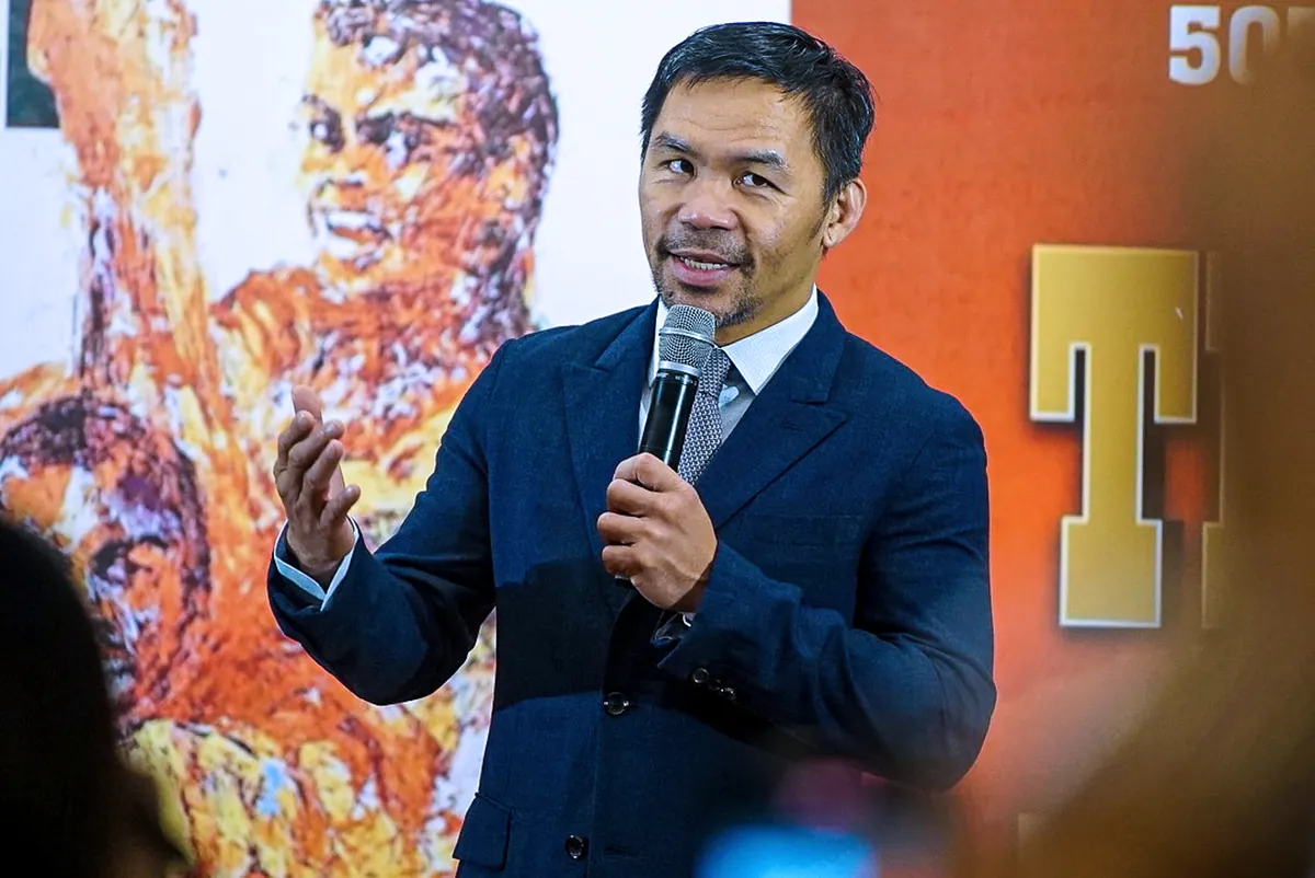 Manny Pacquiao in a navy suit speaking into a microphone in front of a boxing mural
